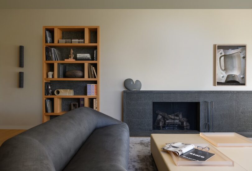 A modern living room with a gray sofa, a light coffee table with books, a black fireplace, a wooden bookshelf, and minimal decorative objects and artwork.