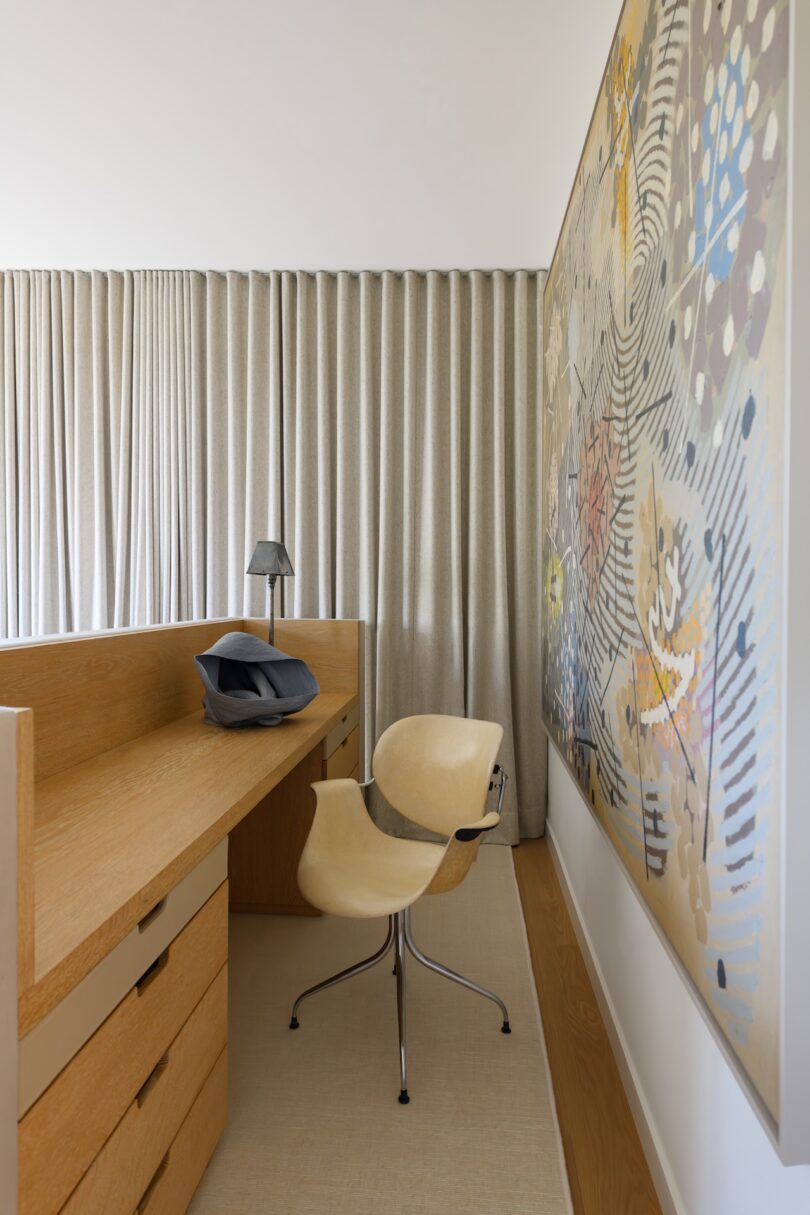 Minimalist home office with a light wood desk, modern beige chair, gray lamp, and abstract wall art. Floor-length beige curtains cover the window in the background.