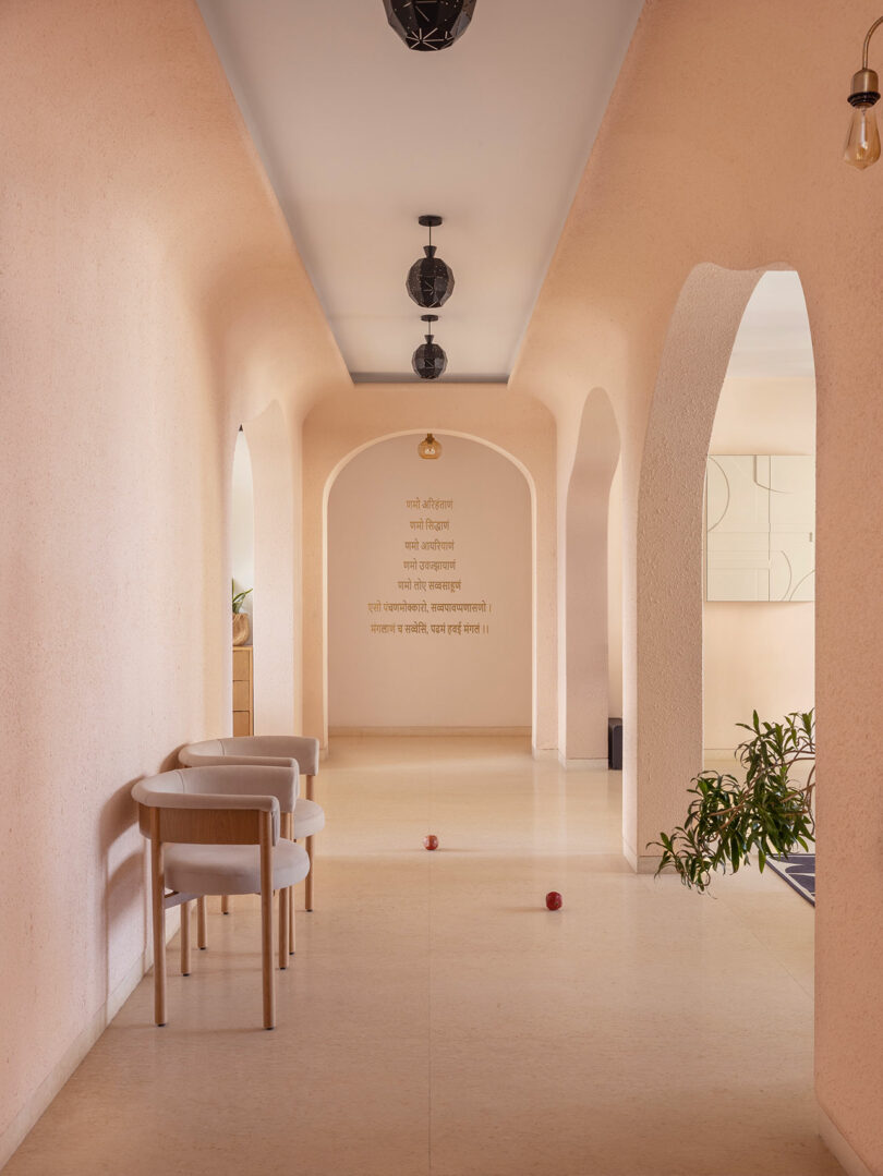 A hallway with arched doorways, light peach walls, two chairs, and red balls on the floor. Hindi text is written on the far wall, and a plant is visible in the background.