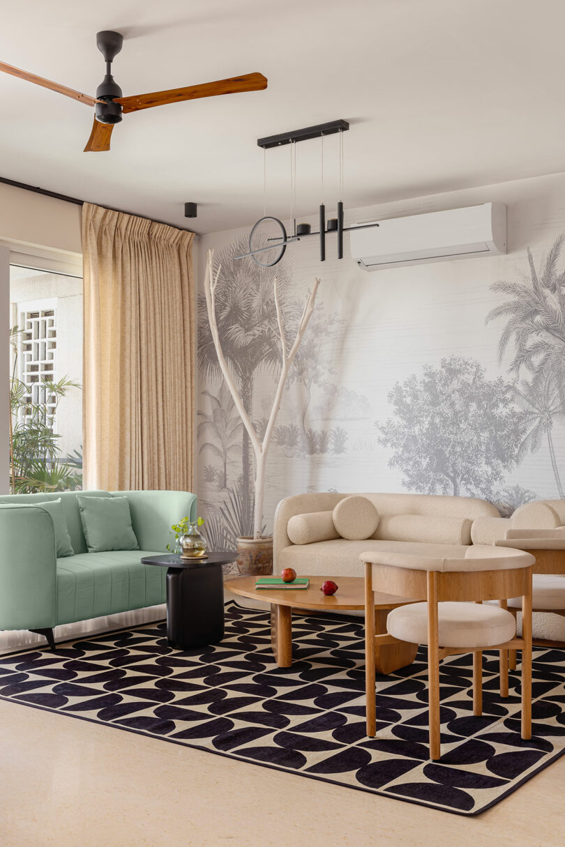 Modern living room with light-colored furniture, a patterned black-and-white rug, ceiling fan, wall-mounted air conditioner, and a nature-themed mural on the wall.