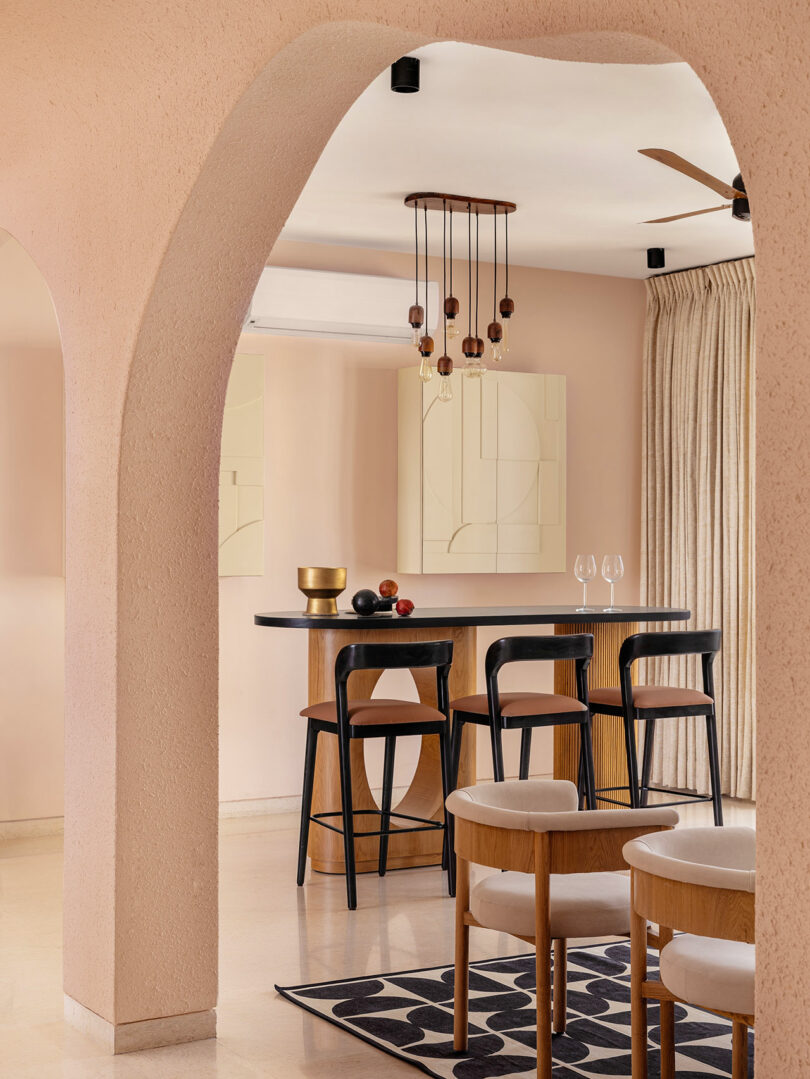 A modern dining area with black chairs and a bar-height table, pendant lights above, neutral walls, and archways, viewed through an arched entryway.